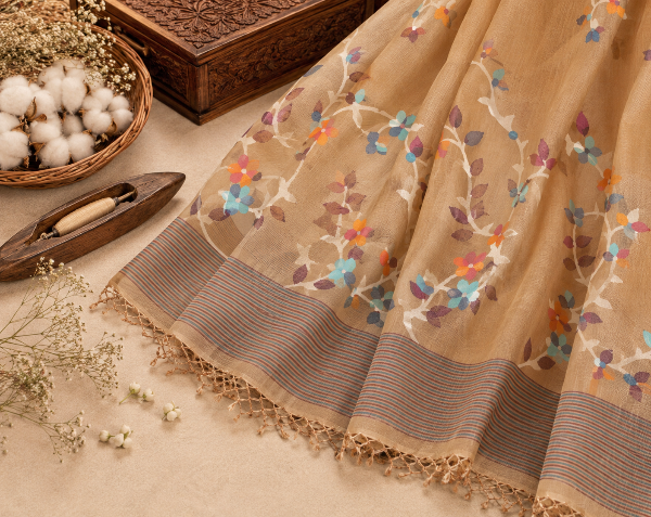 A beige sheer saree or fabric draped on a surface, decorated with colorful floral embroidery in blue, orange, purple, and white. The fabric features a striped border in brown and beige tones with a delicate lace edge.